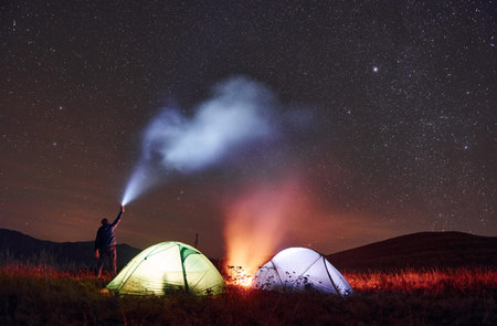 Man holds device with light beam. Two iluminated tents with campfire under stars at mountains at...