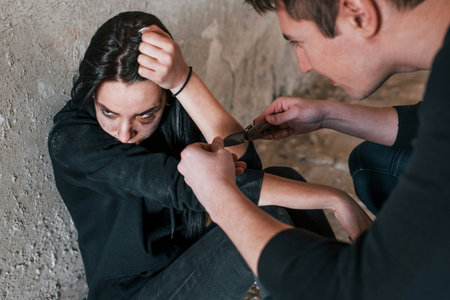 Kidnapped woman sitting and threatened by man with knife in hands in abandoned buildingの写真素材