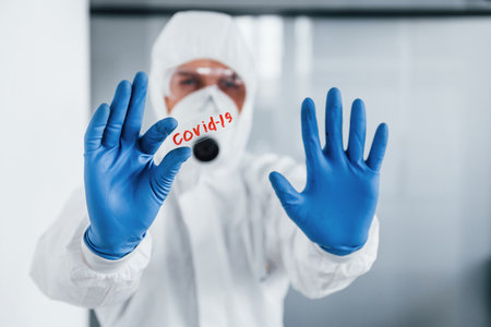 Male doctor scientist in lab coat, defensive eyewear and mask holds glass with covid-19 word on itの写真素材