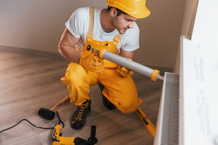 Handyman in yellow uniform works indoors with heat battery by using special tool. House renovation...