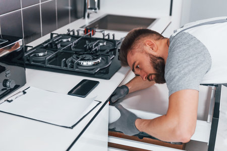 Young professional plumber in grey uniform working on the kitchenの写真素材
