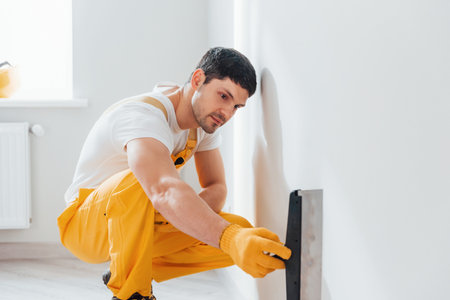 Handyman in yellow uniform polishing wall indoors. House renovation conceptionの写真素材