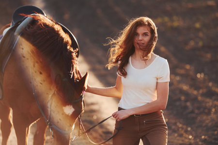 Young woman in protective hat walking with her horse in agriculture field at sunny daytimeの写真素材