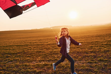 Happy little girl running with kite in hands on the beautiful field at sunrishe timeの写真素材