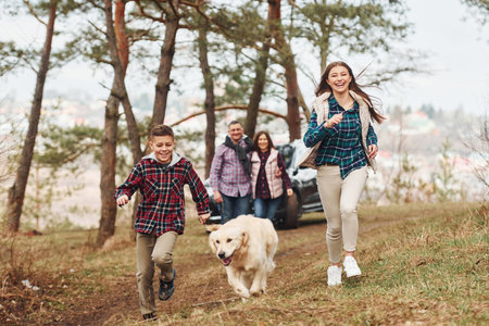 Happy family runs and having fun with their dog near modern car outdoors in forestの写真素材