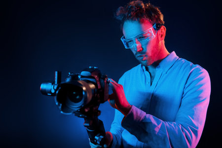 Holds camera. Neon lighting. Young european man is in the dark studioの写真素材