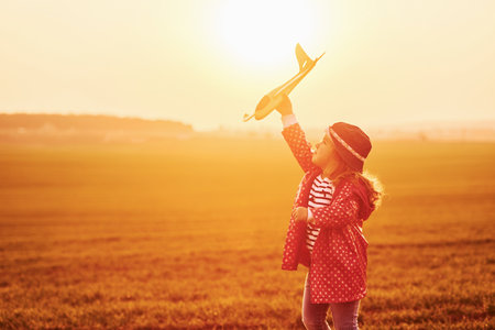 Illuminated by orange colored sunlight. Cute little girl have fun with toy plane on the beautiful field at daytimeの写真素材