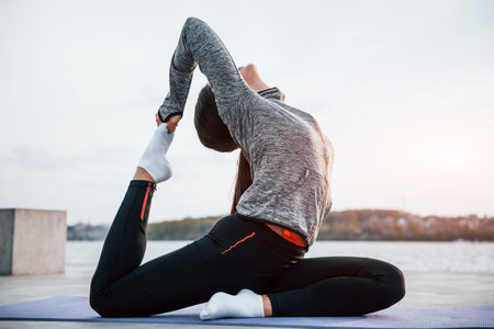Young sportive girl doing yoga exercises on fitness mat outdoors near lake at daytimeの写真素材