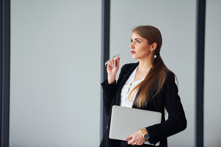 Standing against grey background. Young adult woman in formal clothes is indoors in the officeの写真素材