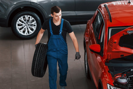 Worker in black and blue uniform holds car wheel and walks with in near red automobileの写真素材