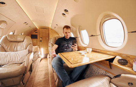 Passenger in casual clothes sits by table with phone in hands. In airplane