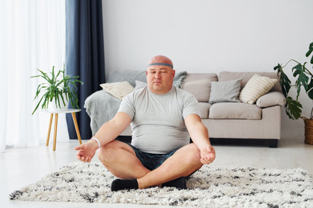Doing yoga exercises. Funny overweight man in casual clothes is indoors at homeの写真素材