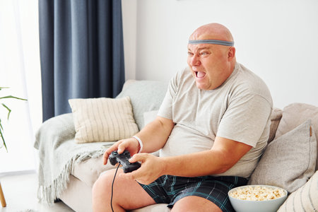 Plays video game. Funny overweight man in casual clothes is indoors at home