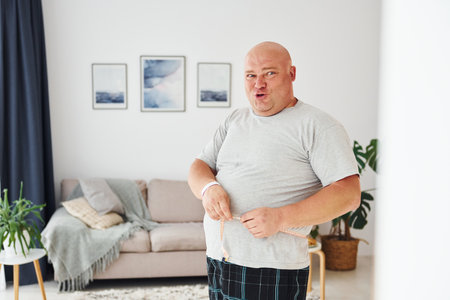 Measuring waist. Funny overweight man in casual clothes is indoors at homeの写真素材