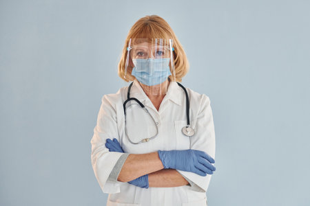 Senior female doctor in white coat is standing indoors against blue backgroundの写真素材