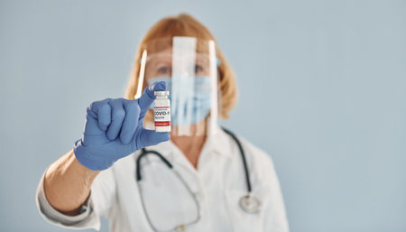 Senior female doctor in white coat is standing indoors against blue backgroundの写真素材