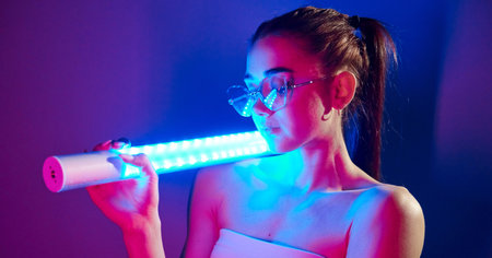 Holds neon lighting stick. Fashionable young woman standing in the studioの写真素材