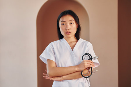 Doctor with stethoscope. Young serious asian woman standing indoorsの写真素材