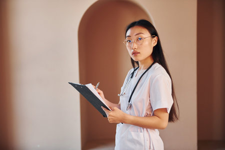 Holds notepad in hands. Young serious asian woman standing indoorsの写真素材