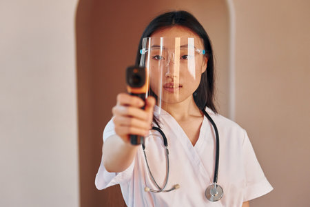 Holds infrared thermometer. Young serious asian woman standing indoorsの写真素材