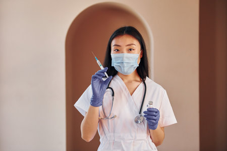 Holds vaccine and syringe. Young serious asian woman standing indoorsの写真素材