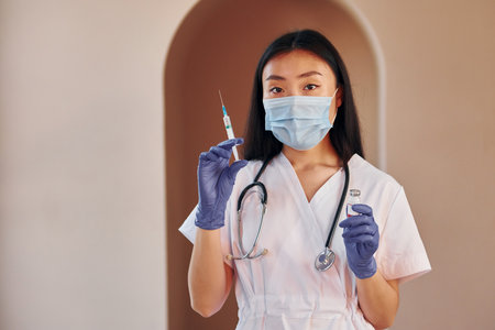 Holds vaccine and syringe. Young serious asian woman standing indoorsの写真素材