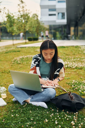 On the grass with laptop on legs. Young asian woman is outdoors at daytimeの写真素材