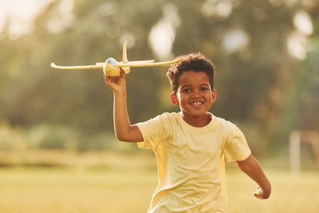 Yellow plane in hands. African american kid have fun in the field at summer daytimeの写真素材