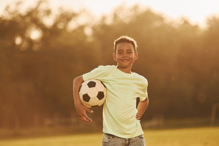 Sits with soccer ball. African american kid have fun in the field at summer daytimeの写真素材
