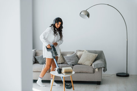 Uses vacuum cleaner. Woman in casual clothes is indoors at homeの写真素材