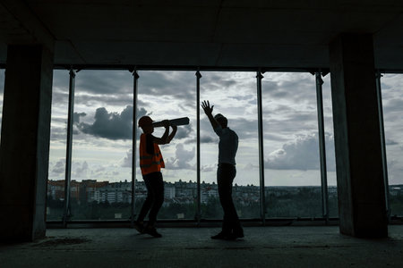 Standing against big window. Man in suit and handyman in orange protective wear is working on...