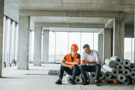 Sitting on the materials. Man in suit and handyman in orange protective wear is working on constructionの写真素材