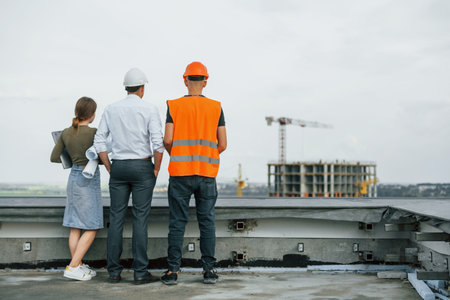 Standing outside with woman. Man in suit and handyman in orange protective wear is working on constructionの写真素材
