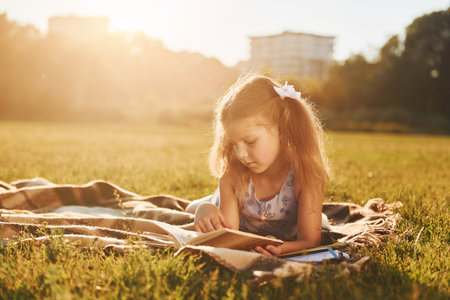 Little girl is reading book while laying down on the grass of field outdoorsの写真素材