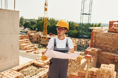 Holding document. Young construction worker in uniform is busy at the unfinished building