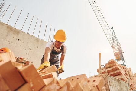 Using bricks. Young construction worker in uniform is busy at the unfinished buildingの写真素材