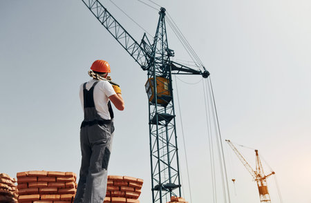 With crane at background. Young construction worker in uniform is busy at the unfinished building