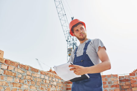 In orange colored hard hat. Young construction worker in uniform is busy at the unfinished building