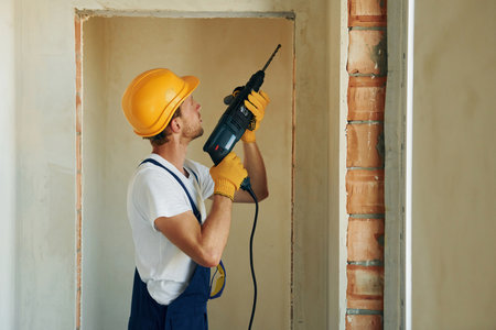 Holding electric drill in hands. Young man working in uniform at construction at daytimeの写真素材