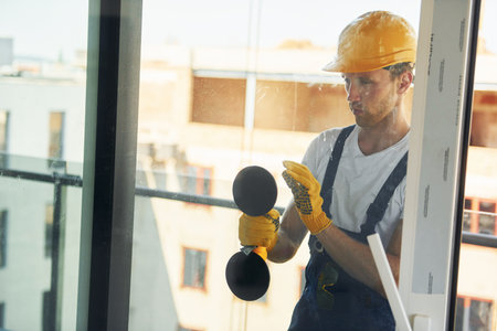 Using tool on glass. Young man working in uniform at construction at daytimeの写真素材