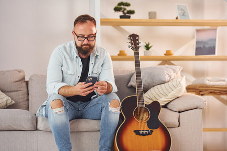 Sitting on the sofa. Man in casual clothes and with acoustic guitar is indoorsの写真素材