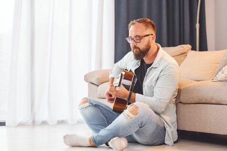 Sitting near the sofa. Man in casual clothes and with acoustic guitar is indoorsの写真素材