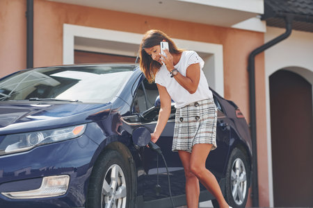 Waiting for the charge of electric car. Woman is outdoors with her modern automobileの写真素材