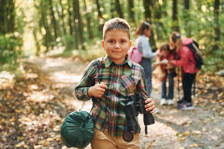 Boy with binoculars standing in front of his friends. Kids in green forest at summer daytime togetherの写真素材