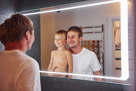 Looking in the mirror in bathroom. Father and son is indoors at home togetherの写真素材