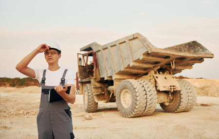 In white hard hat. Worker in professional uniform is on the borrow pit at daytime
