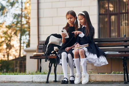 Two schoolgirls is sitting outside together near school buildingの写真素材