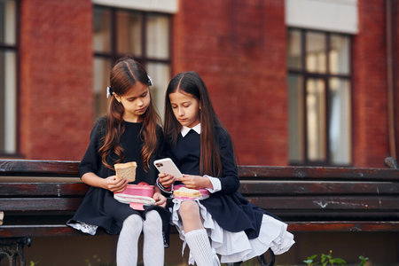 Two schoolgirls is sitting outside together near school buildingの写真素材
