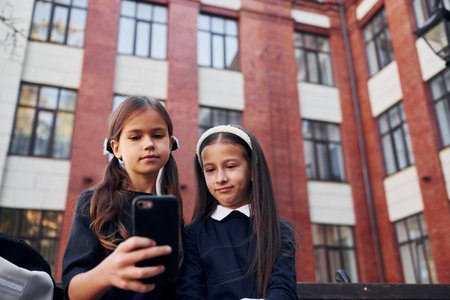 Sitting together. Two schoolgirls is outside near school buildingの写真素材