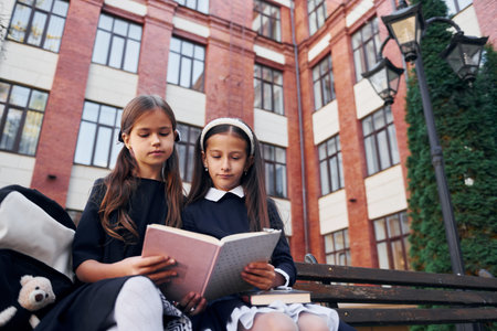 With books. Two schoolgirls is outside together near school buildingの写真素材